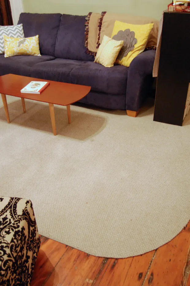 Living room with a purple sofa with yellow and patterned pillows, an orange coffee table with a book, beige round rug, and warm natural light