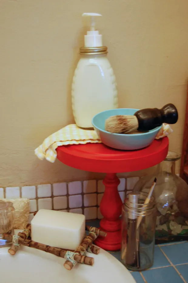 Bathroom vignette with a red pedestal stand holding a lotion pump bottle, teal bowl with shaving brush, soap bar, and glass