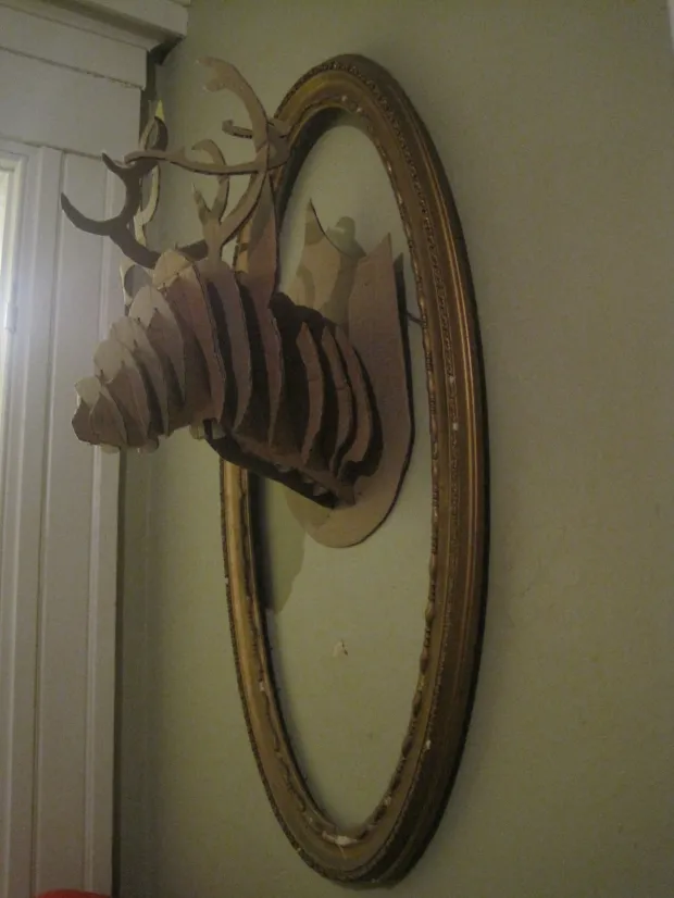 A cardboard cutout deer head sculpture mounted inside an oval gold frame on a wall; brown and gold tones, quirky decor