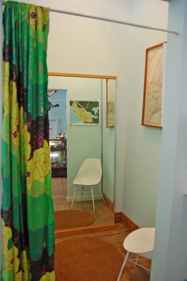 Interior room with a large mirror, framed maps on wall, white chair, wooden floor and green floral curtain, soft pastel tones